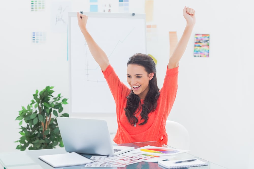 Excited woman raising her arms while working on her laptop in her office Excited woman raising her arms while working on her laptop in her office